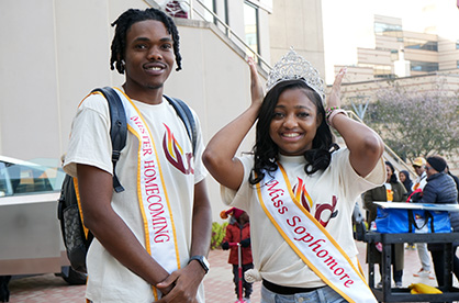 A male and a female student on the UDC plaza. They are wearing royal sashes.