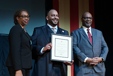 Three people on a stage. The man in the middle is holding a framed award.