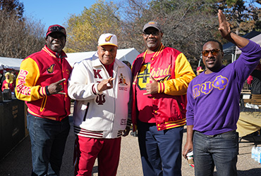 Four men standing outside smiling and making fraternity hand signs.